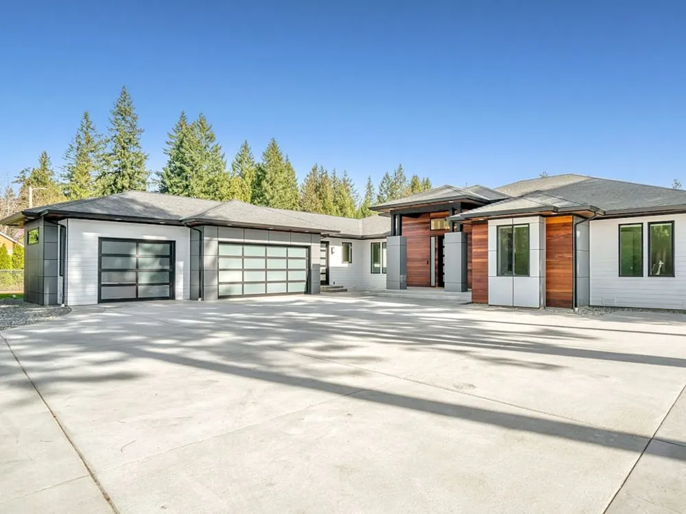 Ranch house with a wide concrete driveway and pine trees behind it